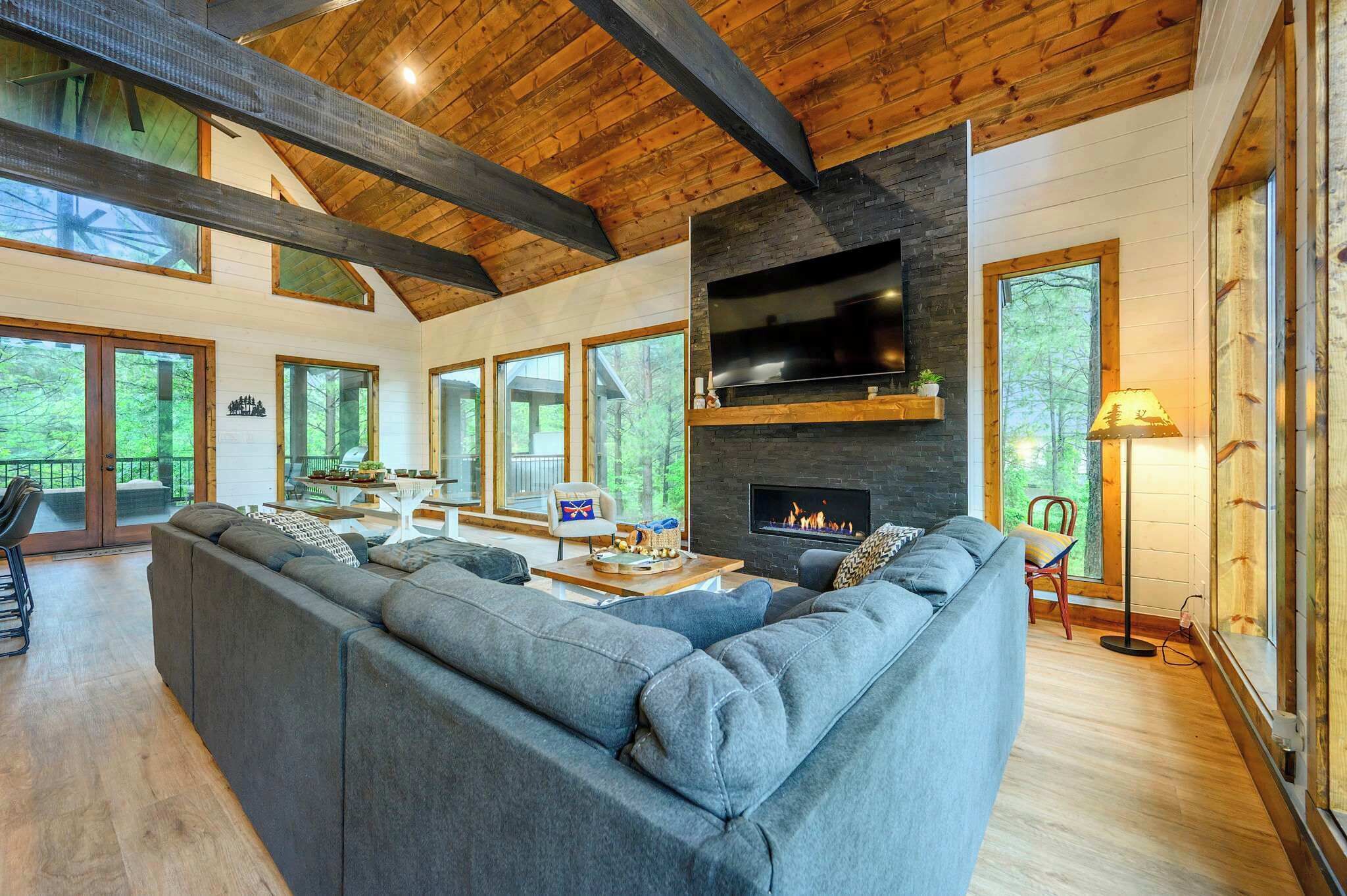 A modern living room with a large sectional sofa, a fireplace, and large windows offering a view of the outdoors.