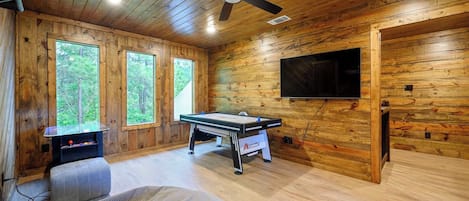 L’intérieur d’un chalet en bois est aménagé avec une table de billard, un ventilateur de plafond, un téléviseur et de grandes fenêtres.