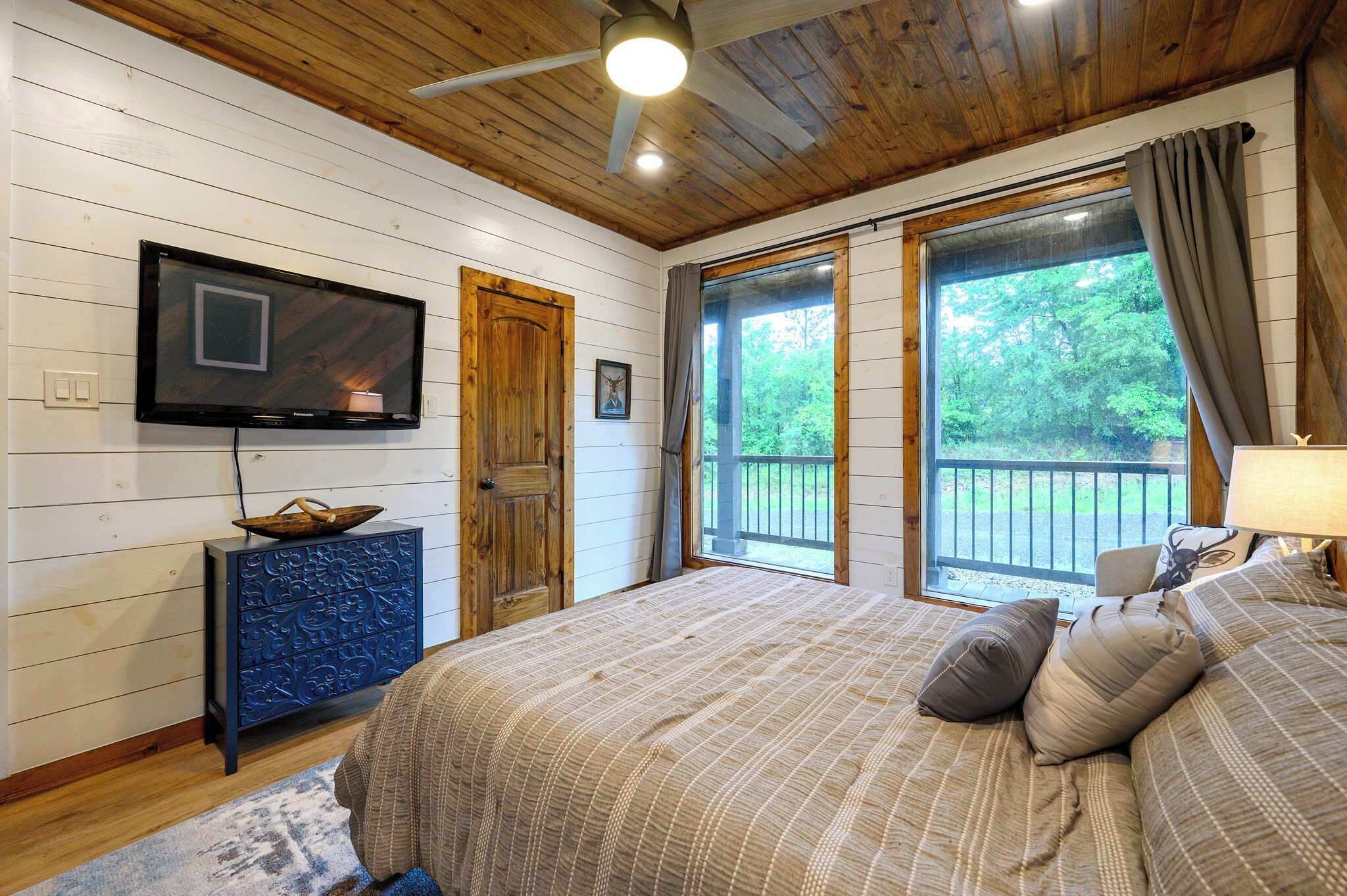 Un dormitorio con cama, televisión, puerta de madera y ventana con vista a follaje.