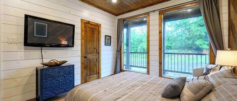 Un dormitorio con cama, televisión, puerta de madera y ventana con vista a follaje.