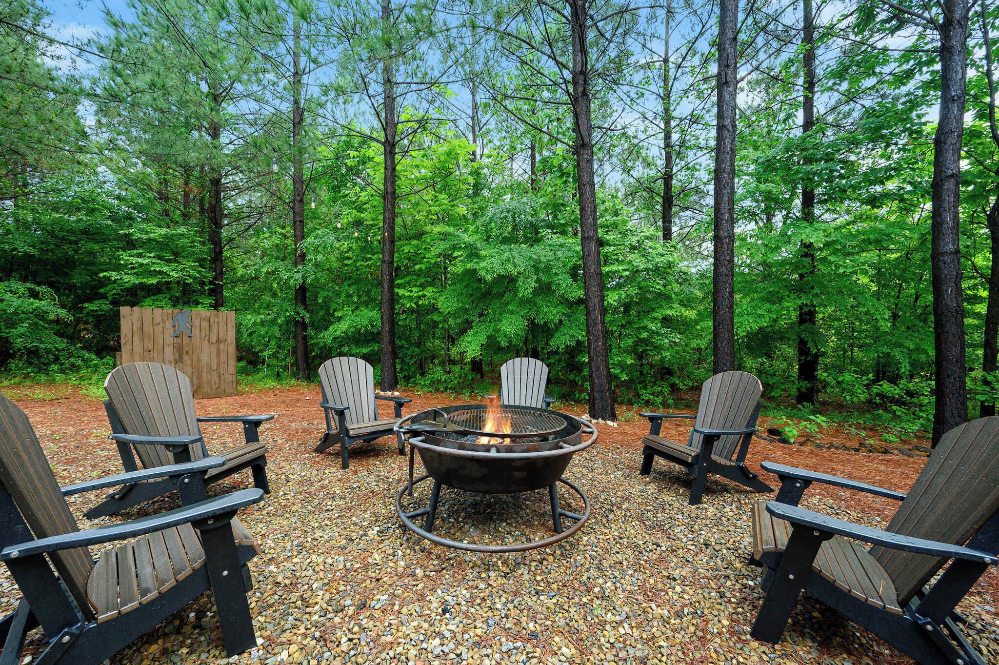 A circular fire pit surrounded by Adirondack chairs in a forested area.