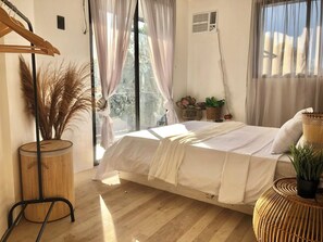 A bedroom with a bed, a window, a plant, a wicker side table, and a wooden floor.