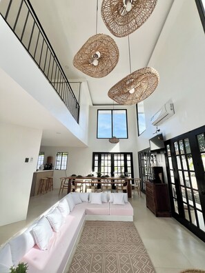 A spacious living area with a high ceiling, a dining table set for four, a sofa with cushions, and a staircase.