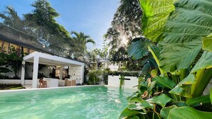 En swimmingpool med ett överbyggt område, omgiven av frodig grönska och en modern byggnad.