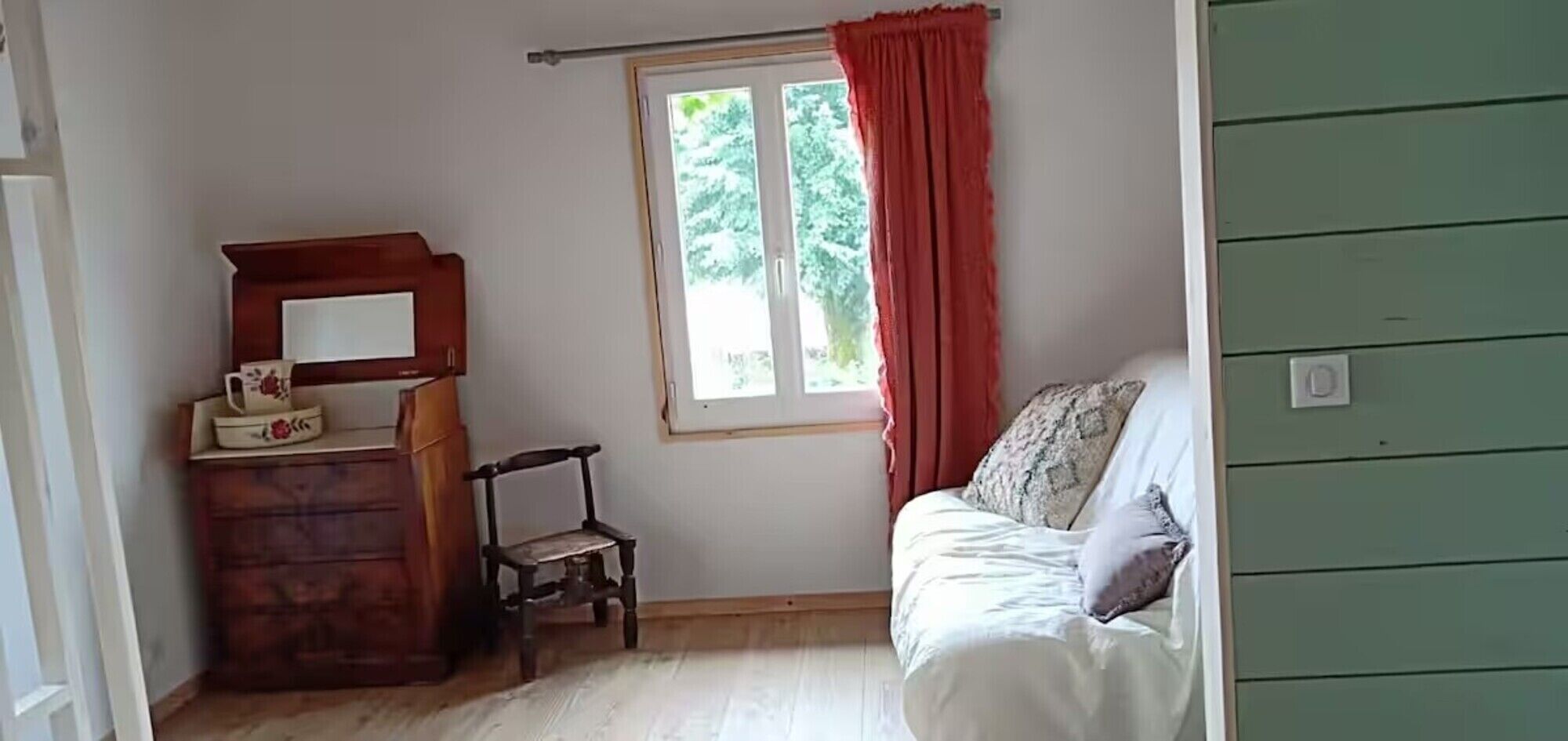 Ein Zimmer mit einem Holzkasten, einem Stuhl, einem Bett mit Kissen, einem Fenster mit Vorhängen und einer grünen Wand.