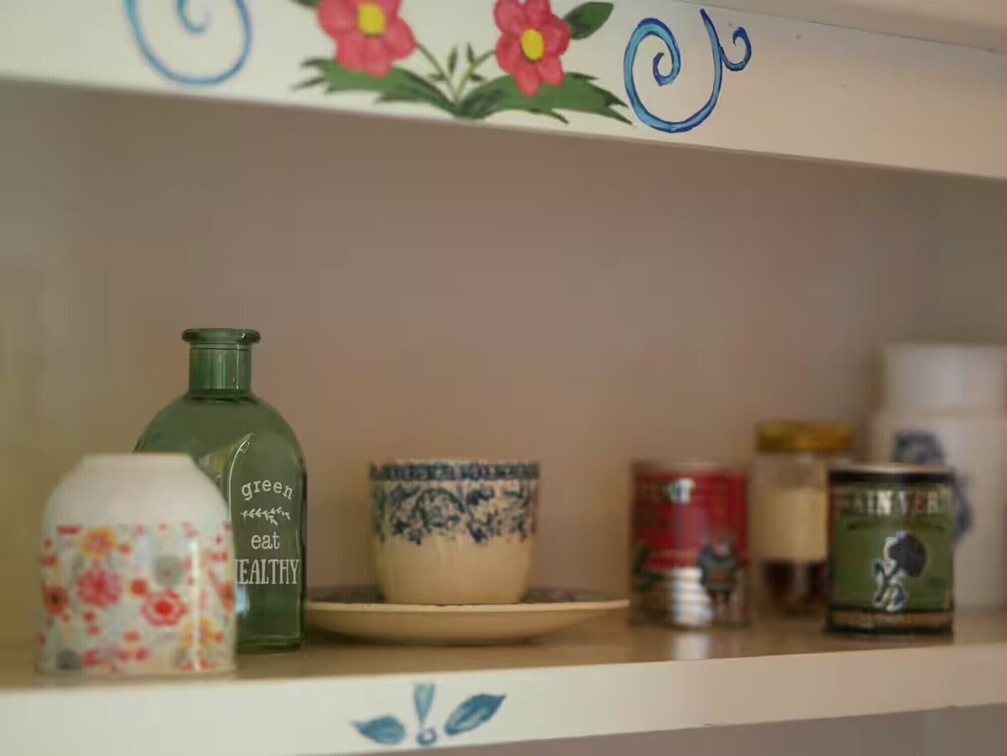 Une étagère avec une bouteille verte portant l’étiquette « vert, excellent et sain », un pot à fleurs, un bol bleu et blanc à motifs, et une boîte de conserve rouge.