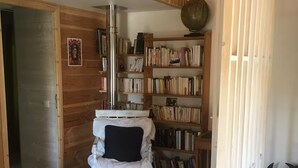 A room with a wooden floor, a white bed with a black pillow, a bookshelf, and a round rug. - Holiday Home “La Voyageuse” with Private Terrace, Garden & Wi-Fi (Gorses)