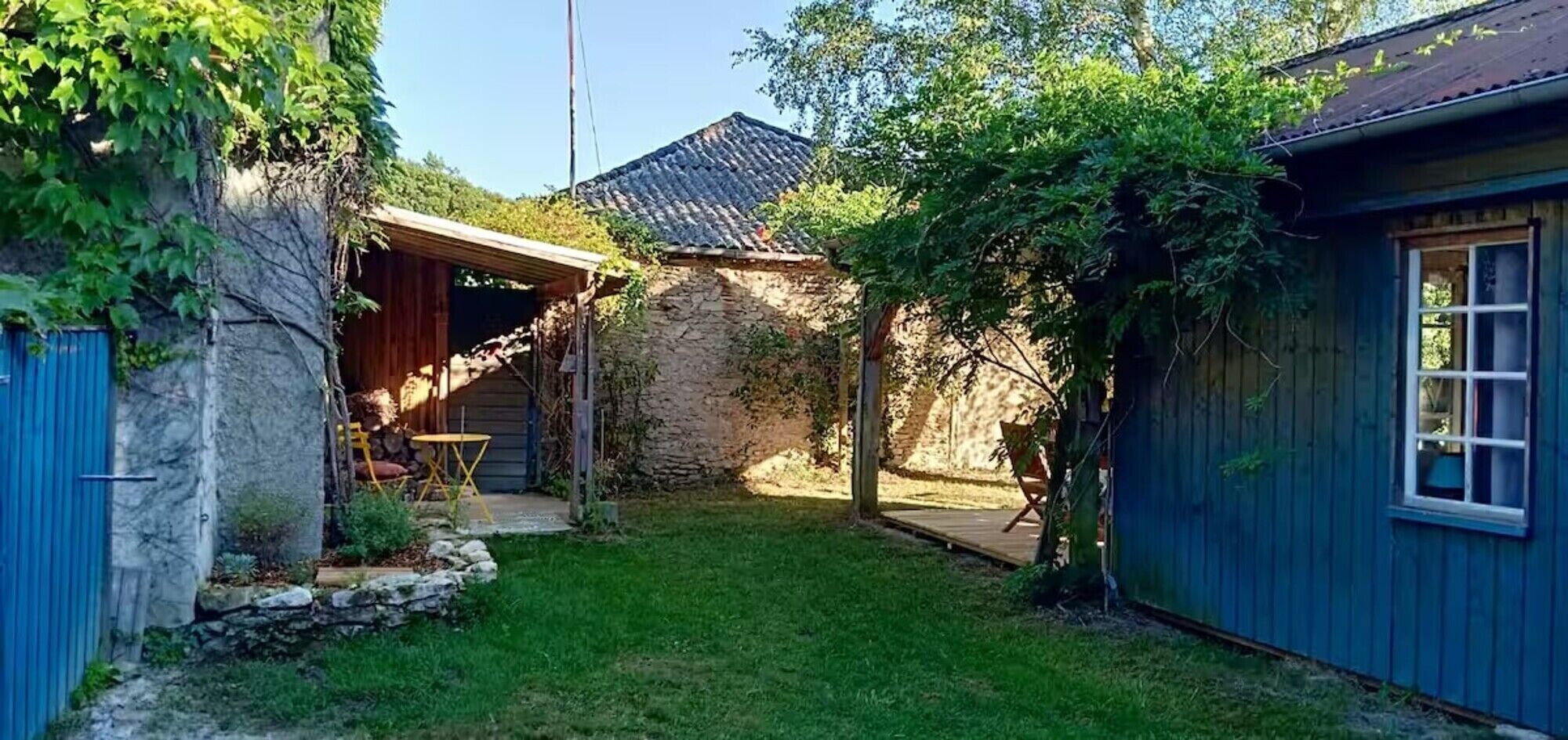 A stone building with a covered patio, a blue shed, and a grassy yard.