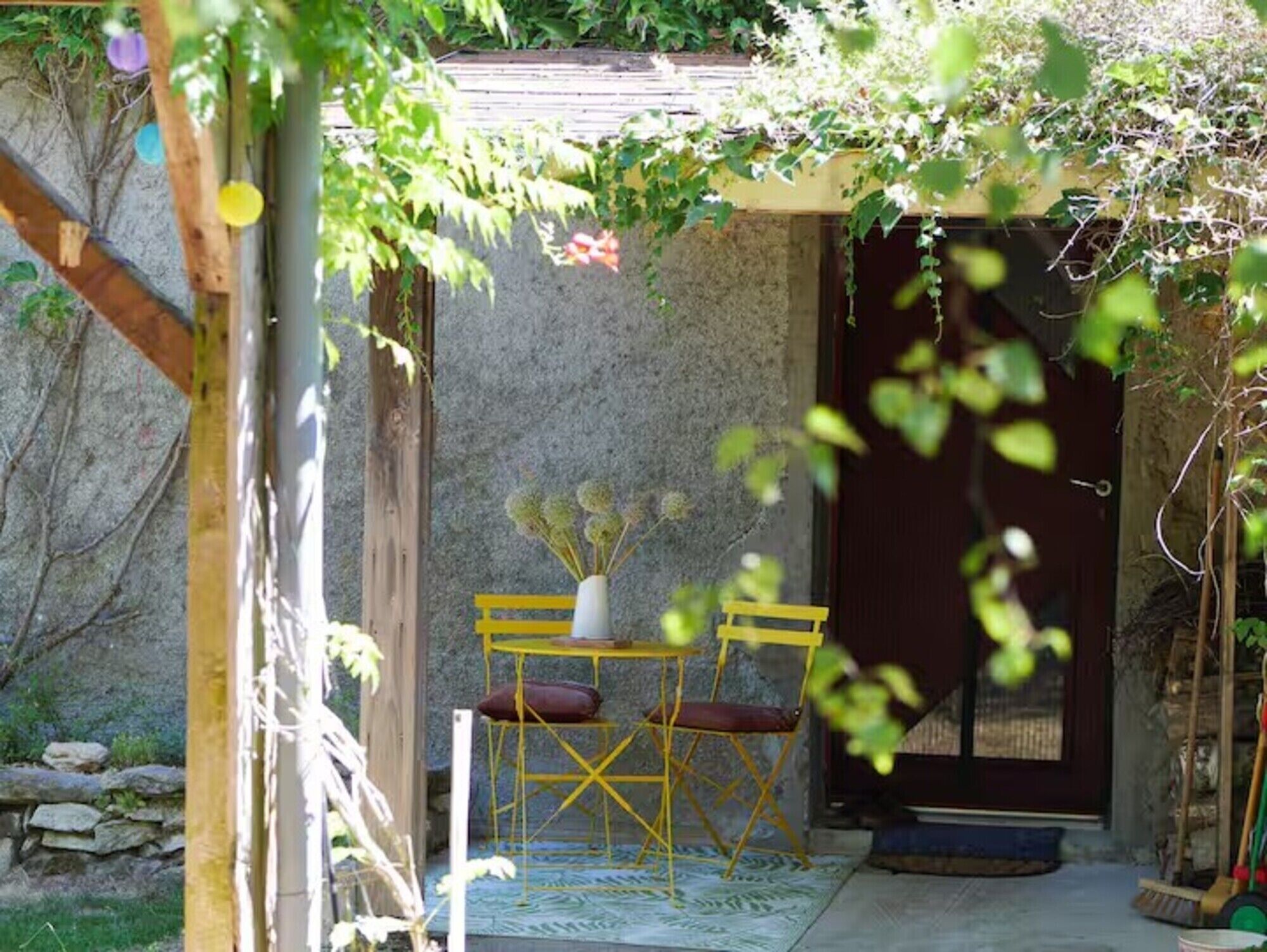Un espace extérieur aménagé dans un style rustique, avec une table et des chaises en bois, un vase rempli de fleurs et un mur en pierre en arrière-plan.
