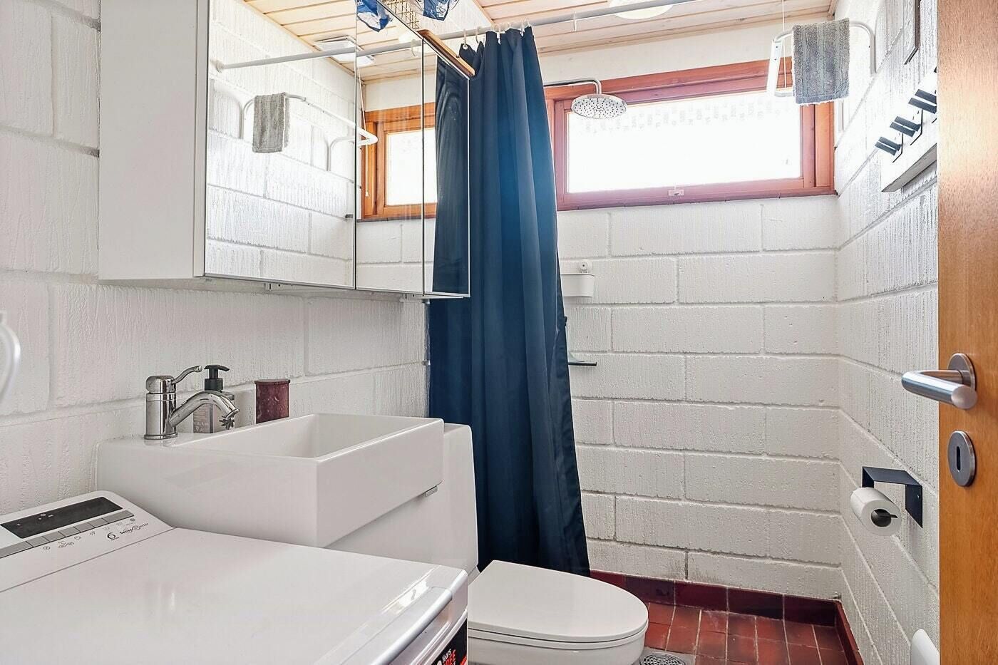 A compact bathroom with a white sink, a toilet, a shower area, and a window with a curtain.