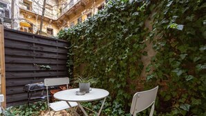 Une petite terrasse extérieure avec une table ronde et deux chaises, entourée d’une clôture en bois et d’un mur recouvert de lierre.