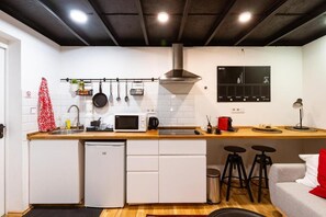 Eine kompakte Küche mit Mikrowelle, Herd und hängenden Kochtöpfen. Es gibt Hocker und einen kleinen Kühlschrank.