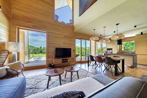 A spacious living area with a large TV, wooden flooring, and a dining area with modern chairs and a kitchen counter.