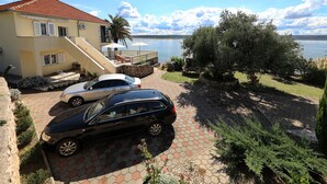 A house with a red-tiled roof, a paved driveway, two cars parked, and a view of a body of water. - Apartment Laura 1 by the Sea (Maslenica)