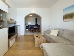 Ein modernes Wohnzimmer mit einer Couch, einem Couchtisch und einem Fernseher. Im Hintergrund ist eine Essecke mit Stühlen und einem Tisch zu sehen.