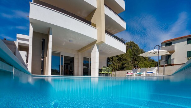 Ein Swimmingpool mit klar blauem Wasser, ein modernes Gebäude mit Balkonen und ein wolkenloser Himmel.