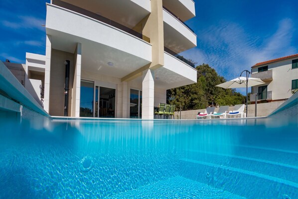 Ein Swimmingpool mit klar blauem Wasser, ein modernes Gebäude mit Balkonen und ein wolkenloser Himmel.