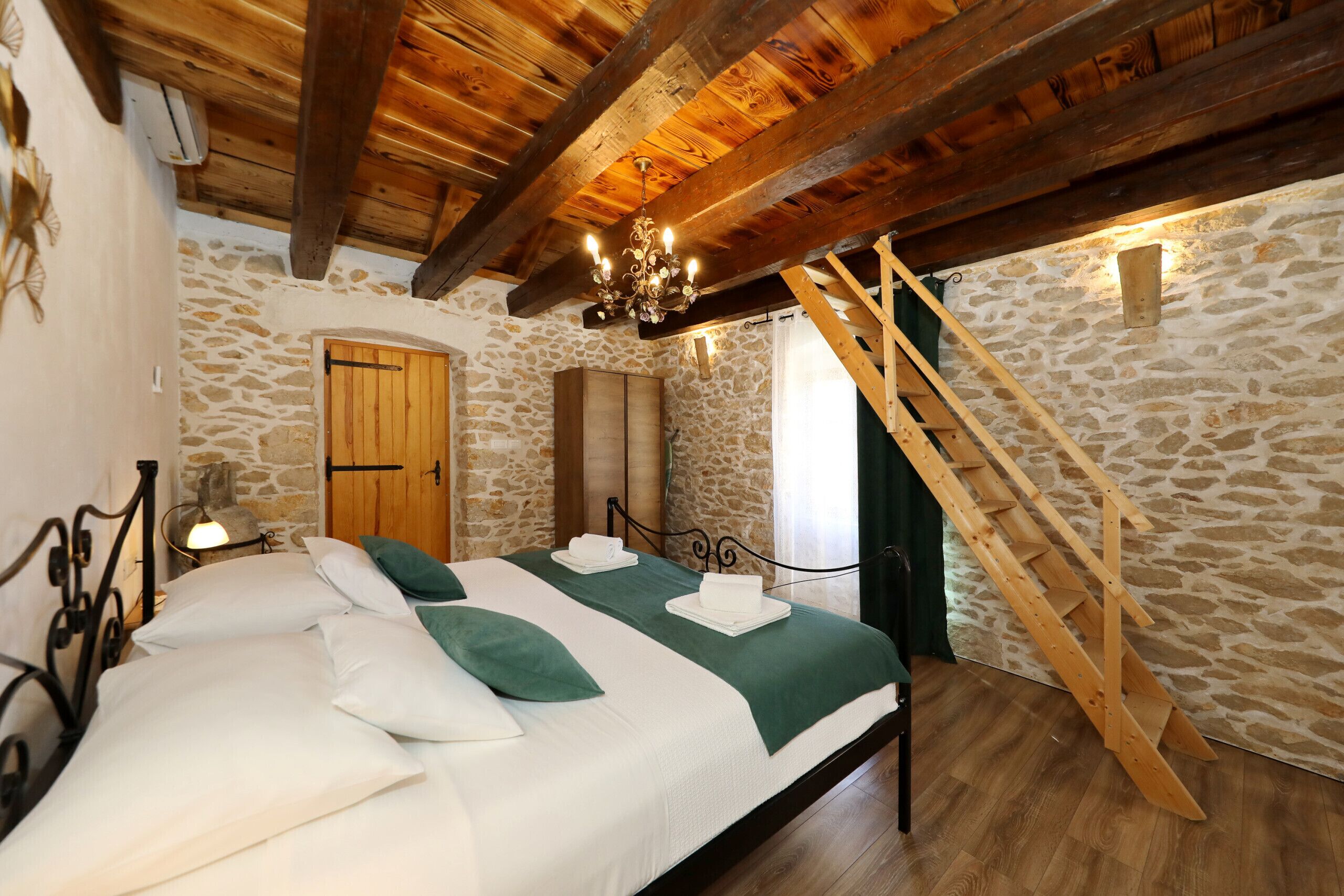 A bedroom with a stone wall, wooden beams, a bed with white and green bedding, a wooden ladder, and a stone fireplace.