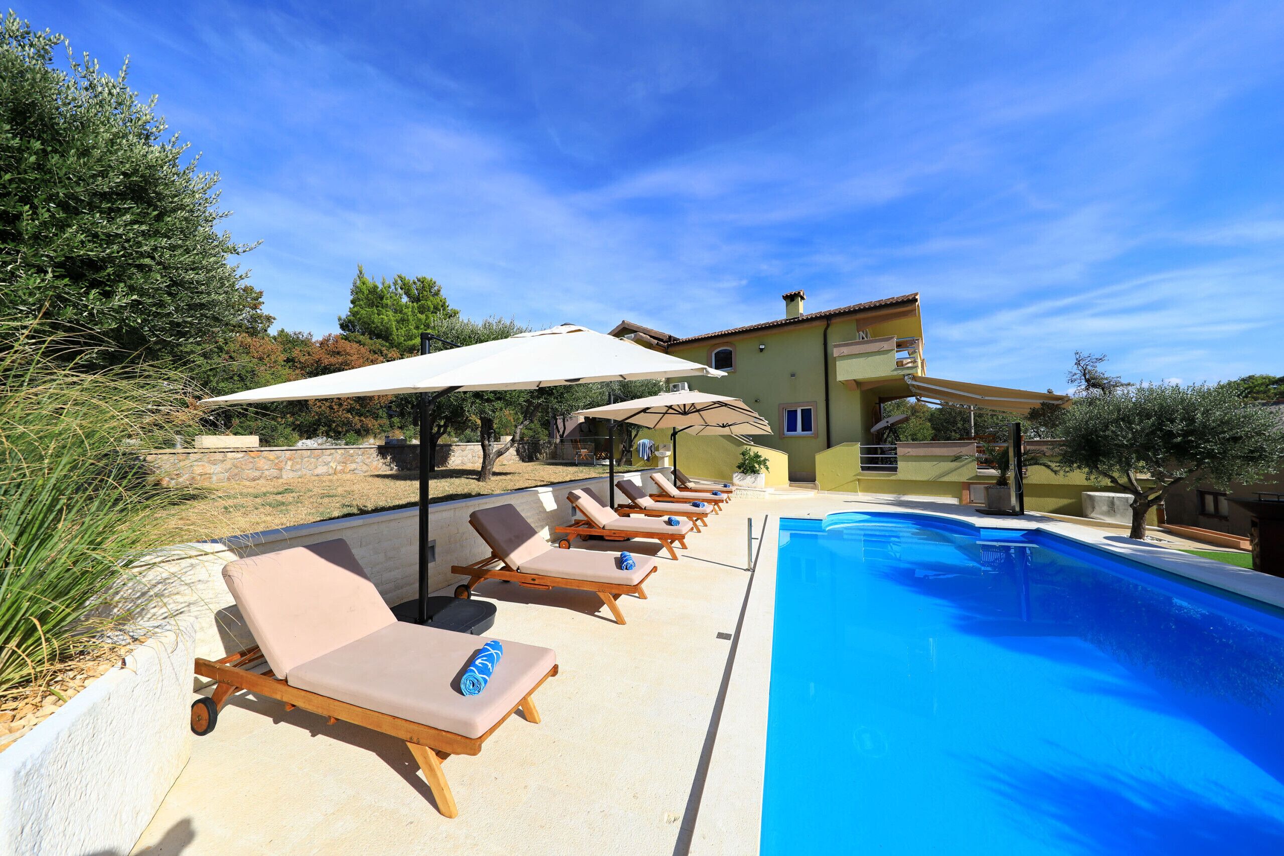 Área da piscina com espreguiçadeiras e guarda-sóis, céu azul e um edifício ao fundo.