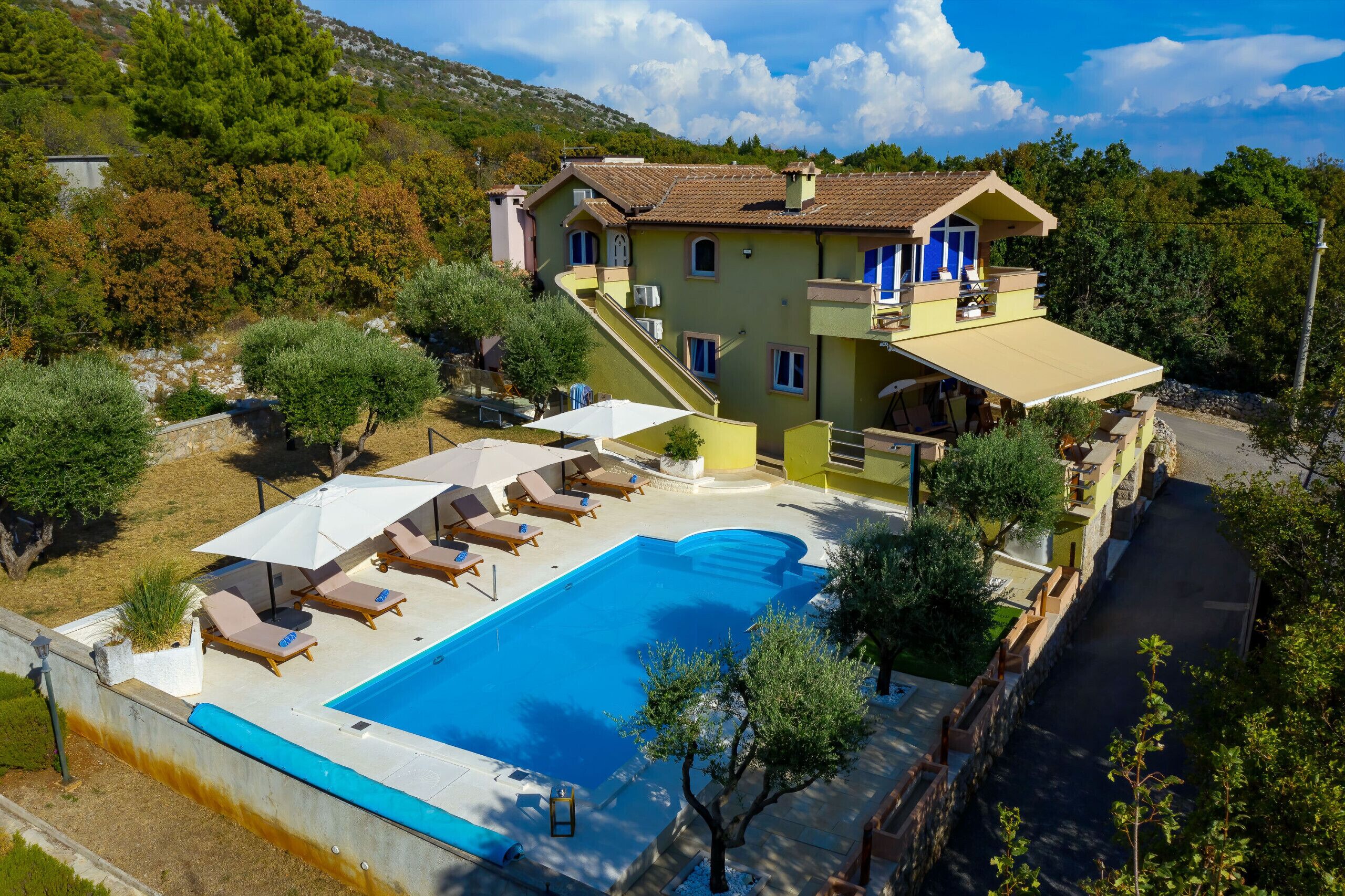 A two-story building with a swimming pool, outdoor seating, and a hillside backdrop.