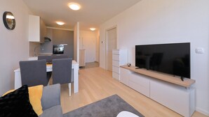 A modern living room with a flat-screen TV, a dining area with chairs and a table, and a kitchen in the background.