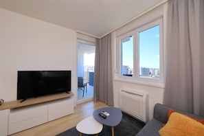 A modern living room with a flat-screen TV, a small table, a sofa, and a large window with a view of buildings. - Apartment Kostela with Balcony and City View (Zadar)