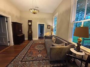 Un salon spacieux avec une grande fenêtre, un tapis à motifs, un canapé, une table avec une lampe et une horloge comtoise.