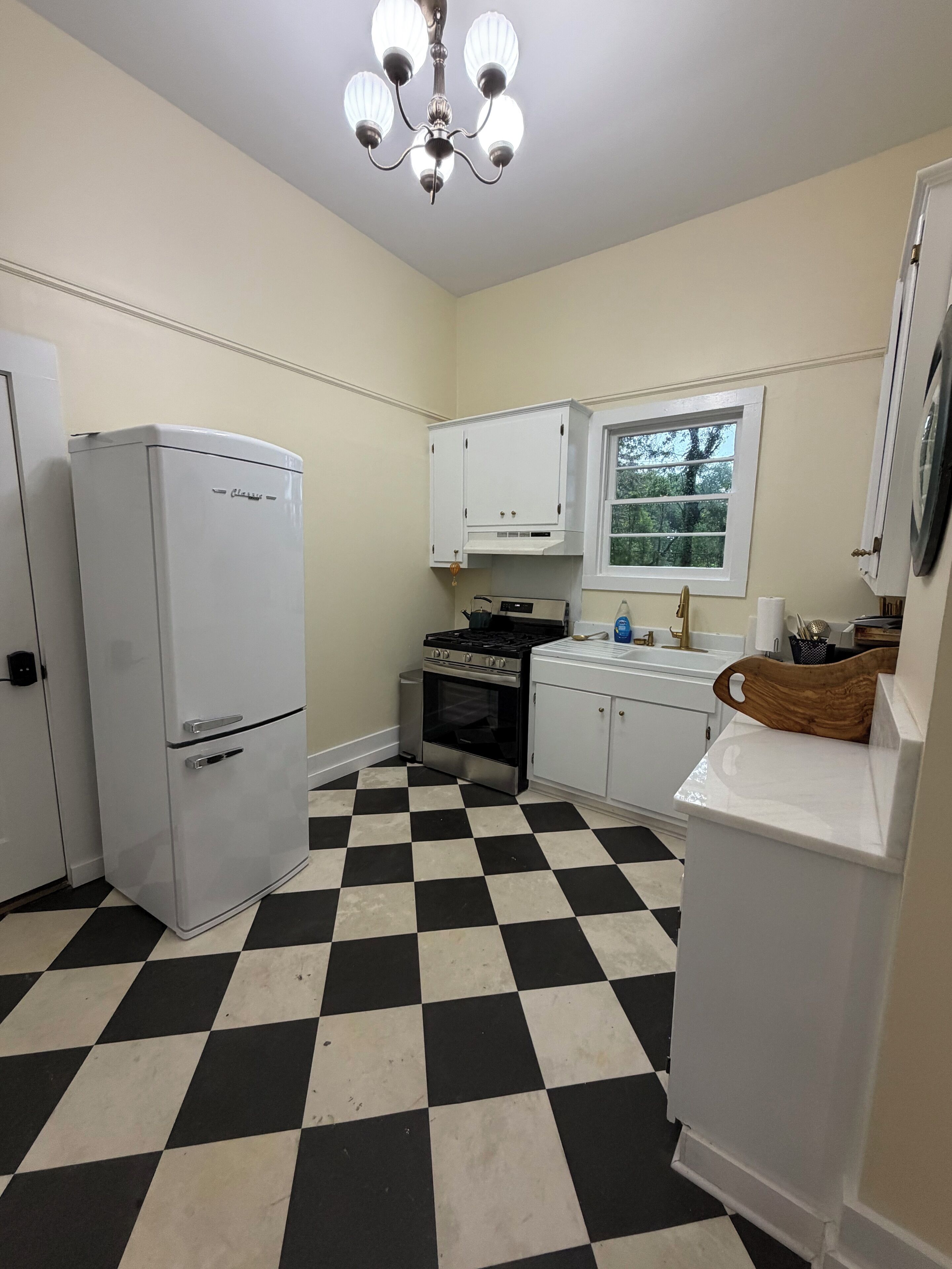Una cocina con piso a cuadros, armarios blancos, un refrigerador, un horno y una araña.