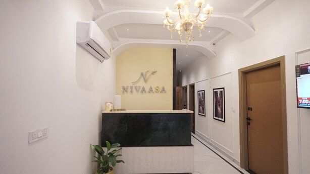 Una mesa de recepción con un cartel que dice 'NIVAASA', una araña, una planta en maceta y una puerta con la etiqueta '002'.