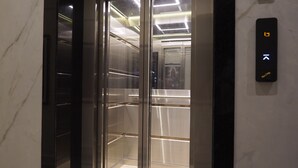 Un ascensor con pared de mármol, puerta de cristal y panel de control con flechas direccionales.