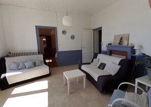 Ein Wohnzimmer mit einem weißen Sofa, einem schwarzen Sofa, einem weißen Couchtisch und einem blauen Sessel. Ein Kamin ist vorhanden, über ihm hängt ein Gemälde.