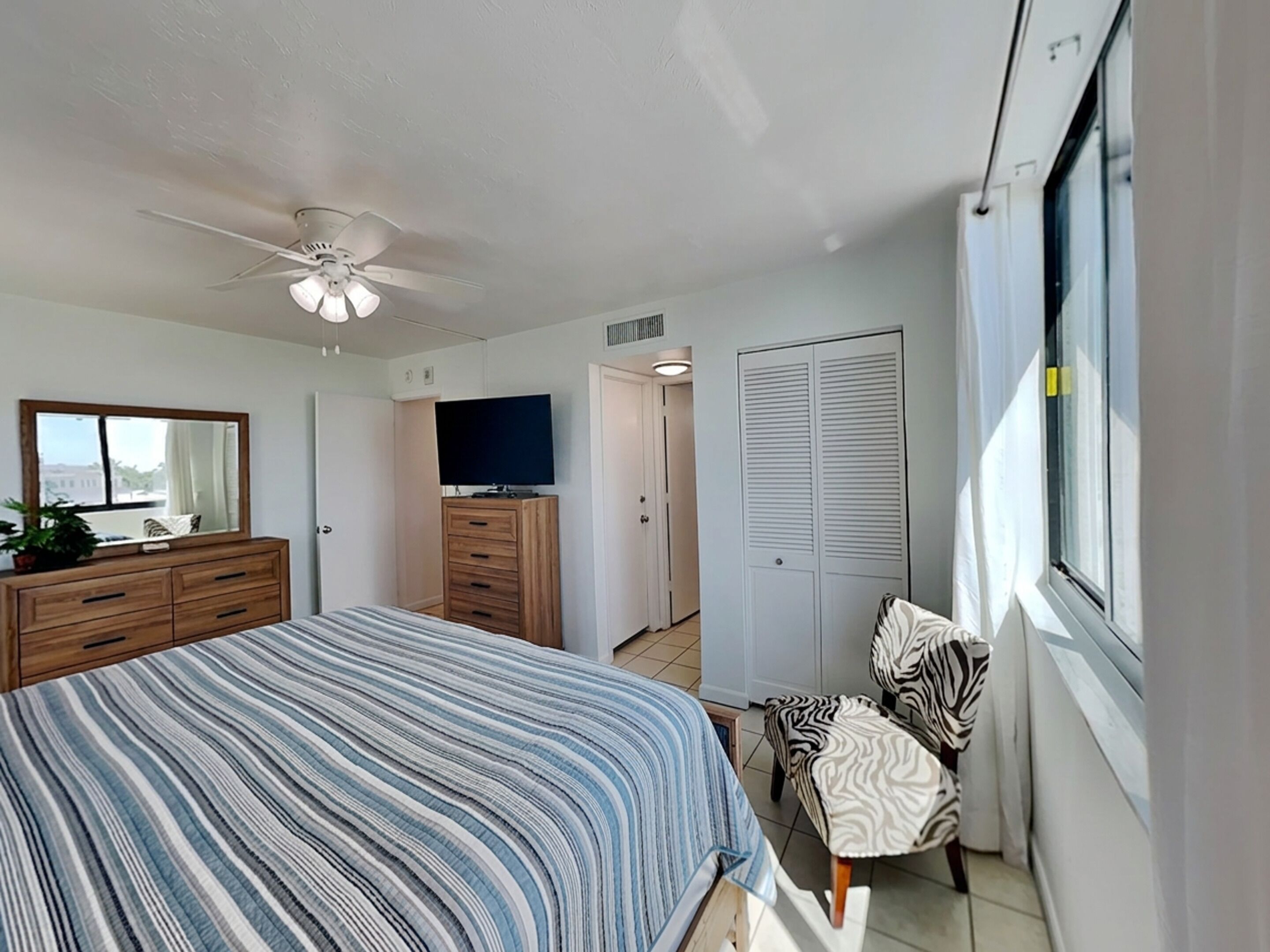 A bedroom with a bed, a ceiling fan, a TV on a wooden cabinet, a wardrobe, a zebra-patterned chair, and a window.