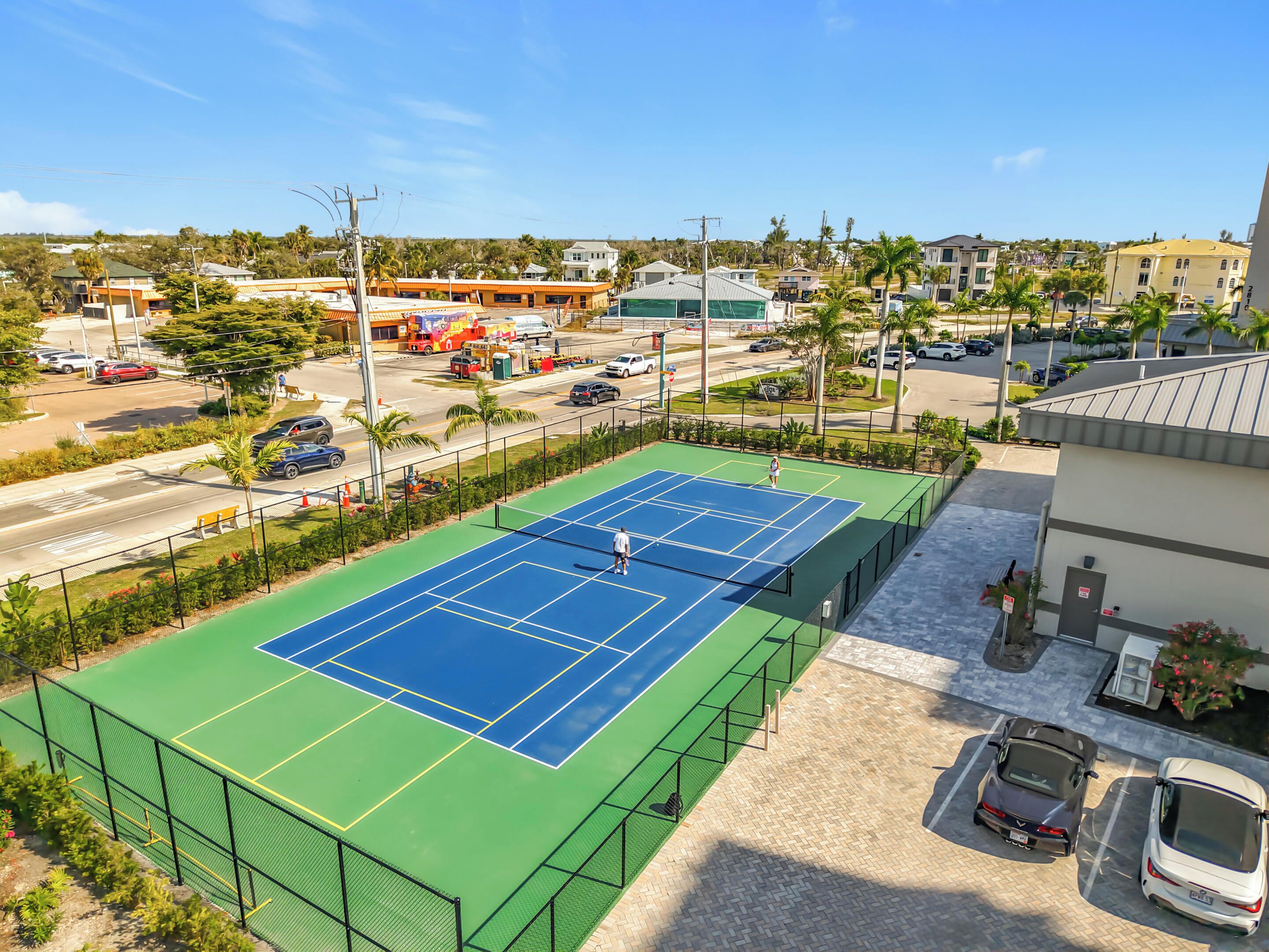 A tennis court with a net, surrounded by a fence, with a parking area and buildings in the background.
