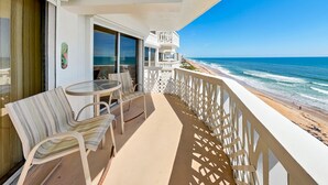 A balcony with a table and chairs, a view of the ocean, and a building with a sign. - Oceanside Retreat 805 | Pet-Friendly + Ocean Views (Ormond Beach)