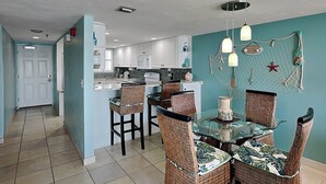 A kitchen with white cabinets, a dining area with wicker chairs and a glass table, and a teal wall with a starfish decoration. - Oceanside Retreat 805 | Pet-Friendly + Ocean Views (Ormond Beach)