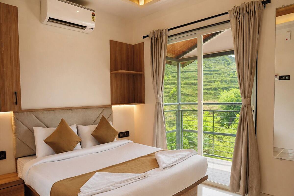 Habitación de hotel con una cama grande, cabecera incorporada, unidad de aire acondicionado, ventana con cortinas y vistas a zonas verdes.