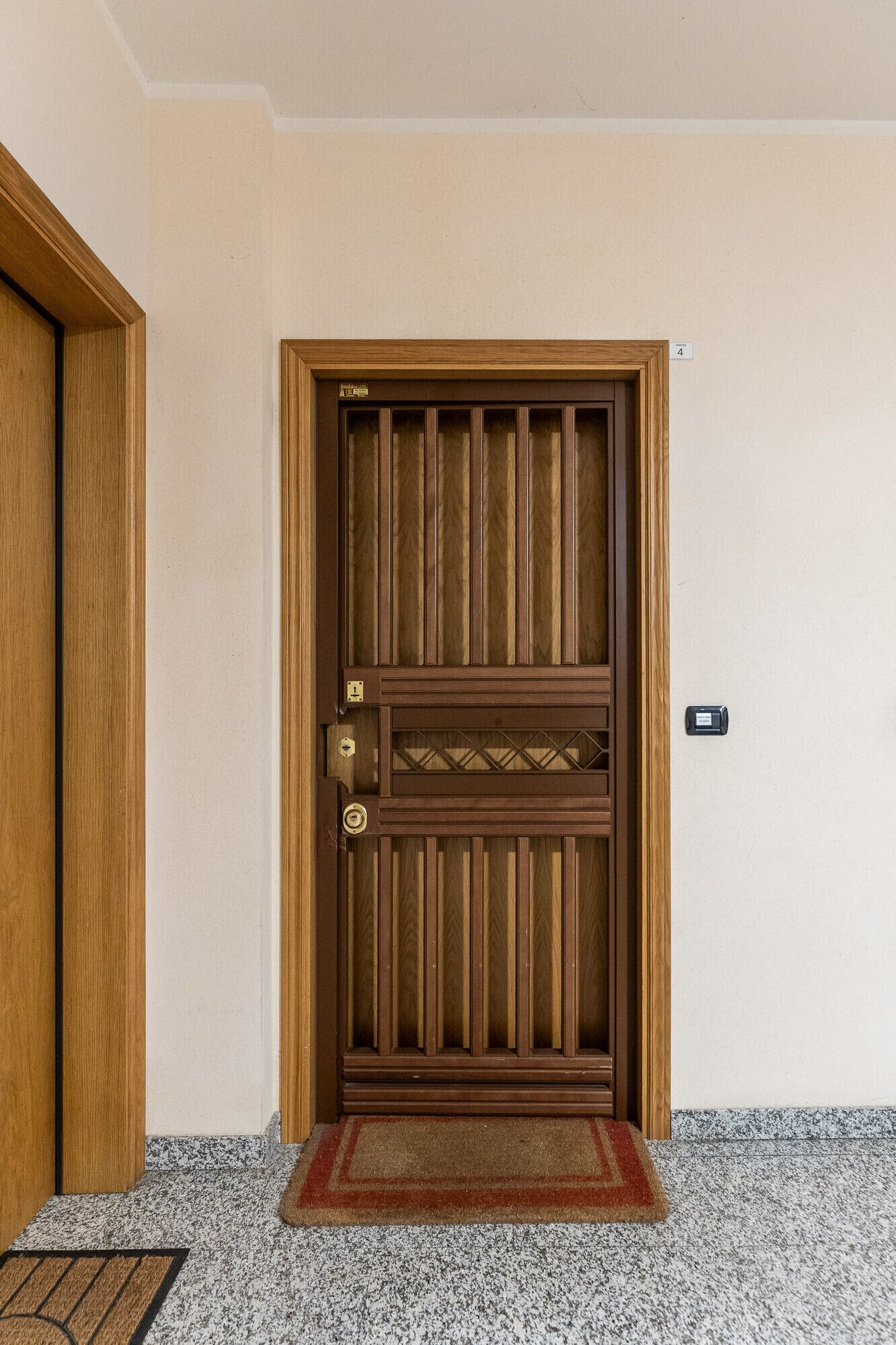A wooden door with a vertical panel design, a doormat, and a wall-mounted device.