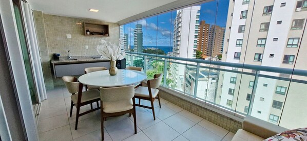 A modern kitchen with a round dining table and chairs, a vase with flowers, and a large window with a city view. - COBAR0150 - Edificio Top House (Salvador)