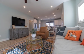 A living room with a sofa, armchair, and coffee table, a television, and a kitchen in the background. - Join us at the Southern Plain Cottage 3-bedroom house close to Auburn campus (Auburn)