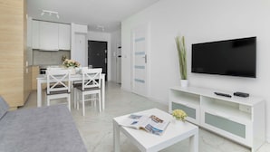 Een moderne woonkamer met een flatscreen televisie, witte meubels, een eethoek en een keuken op de achtergrond.