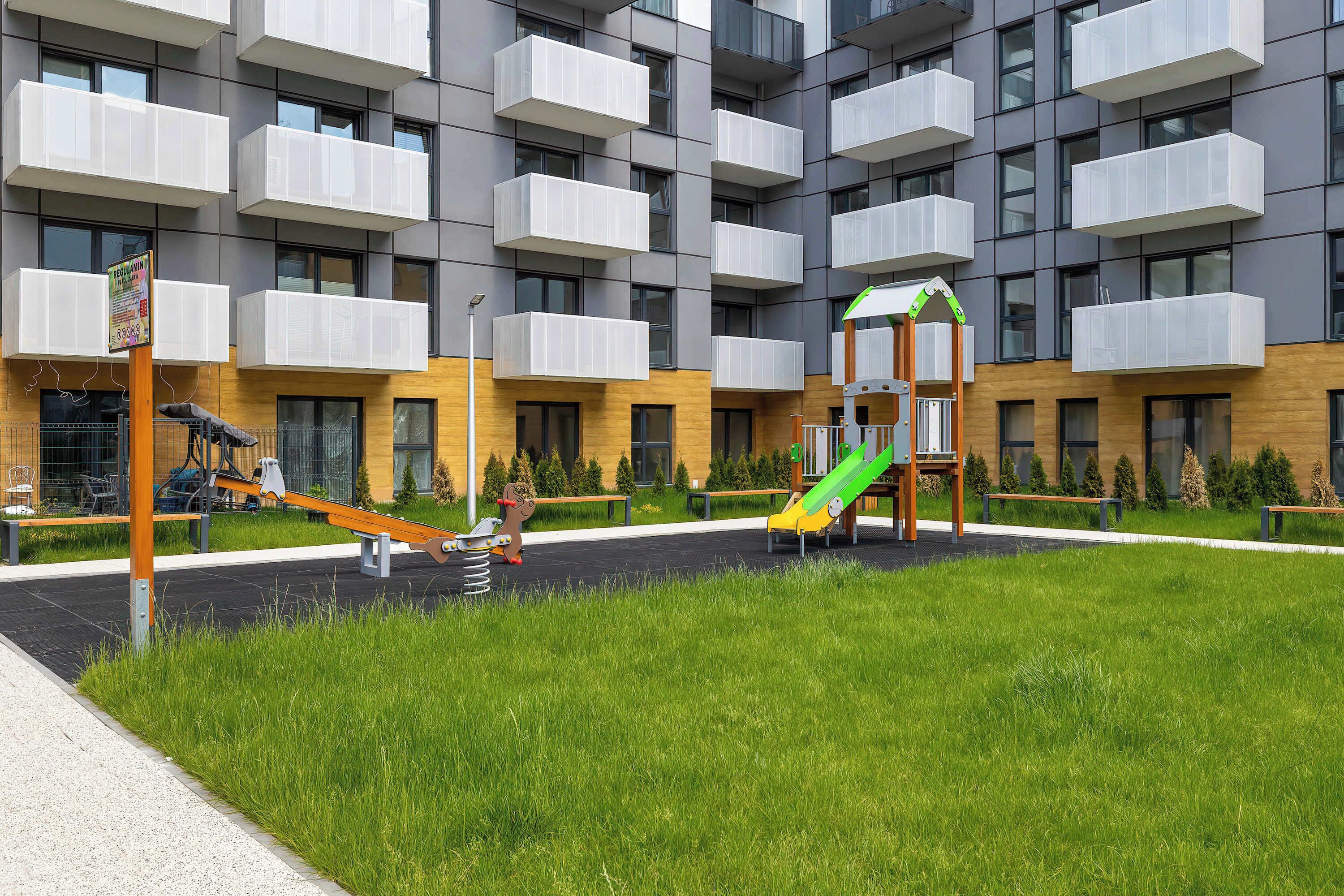 Un parque infantil con columpios, un tobogán, bancos y un edificio con balcones al fondo.