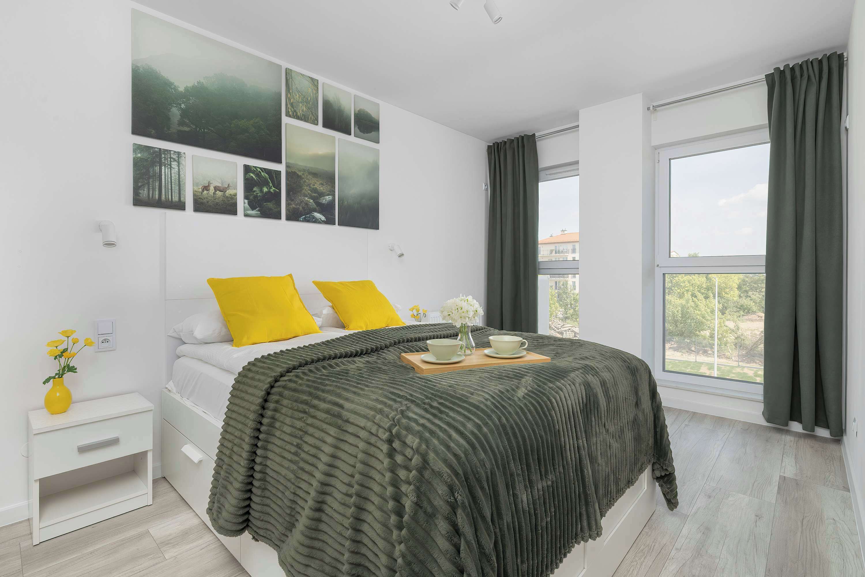 A bright master bedroom features a double bed layered with a textured dark green blanket and bright yellow accent pillows. A collection of nature-themed prints hangs above the headboard, while long green curtains frame the large windows.