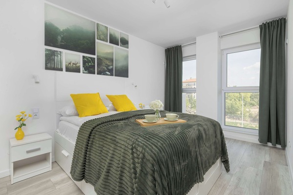 A bright master bedroom features a double bed layered with a textured dark green blanket and bright yellow accent pillows. A collection of nature-themed prints hangs above the headboard, while long green curtains frame the large windows.