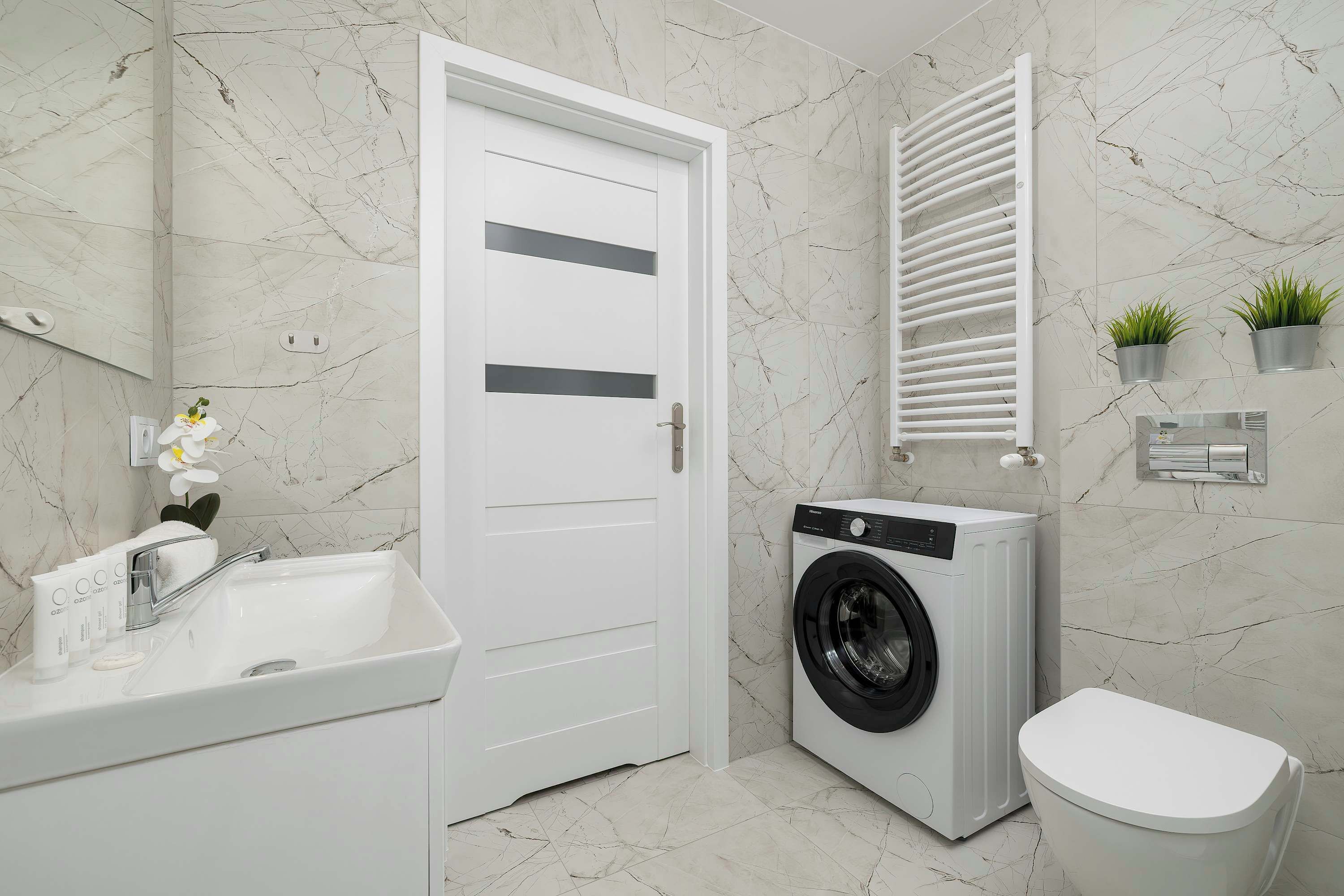 Un baño con puerta blanca, lavadora, inodoro, lavabo y un toallero con plantas.