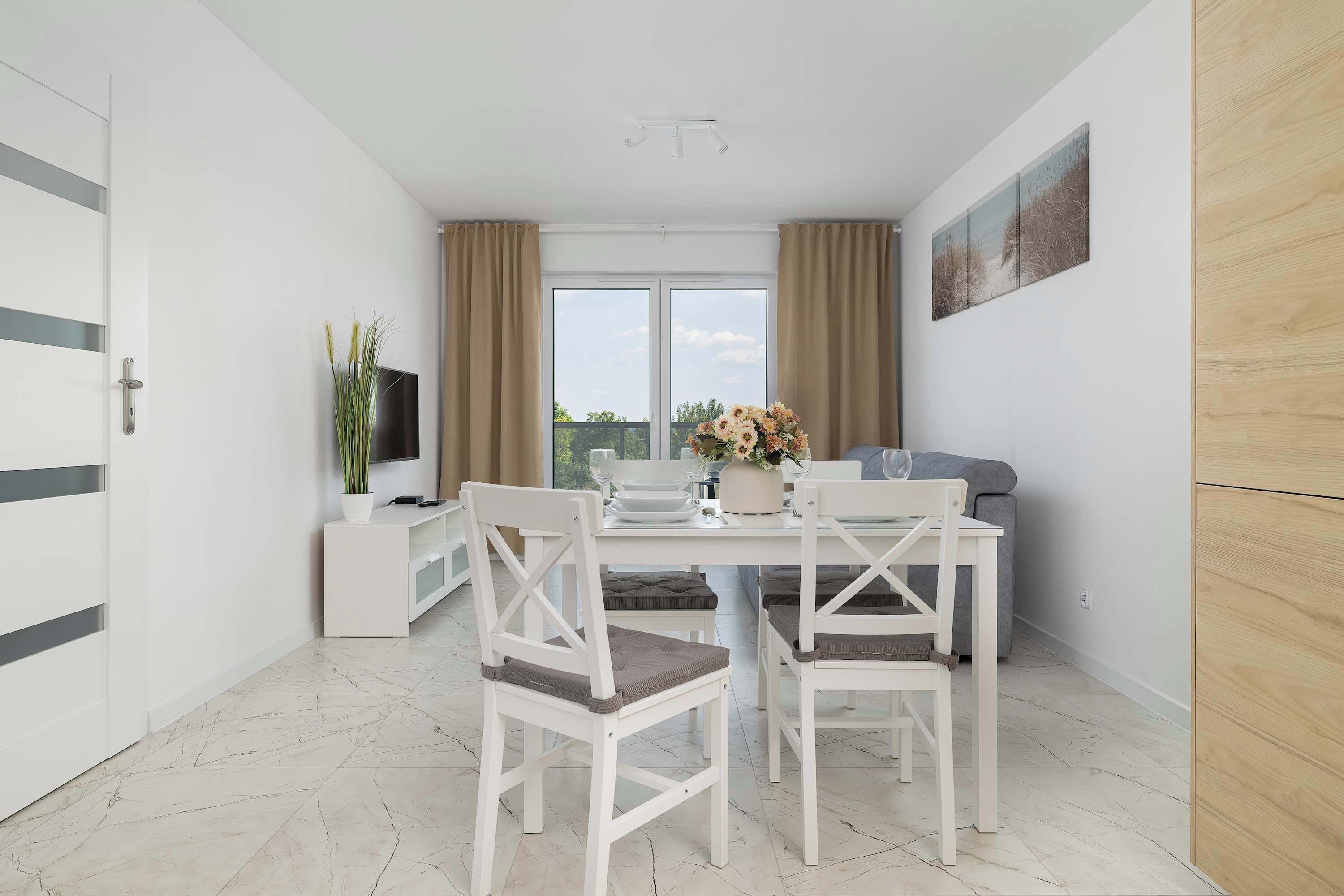 A modern dining area with a white table, two chairs, a vase with flowers, a grey sofa, and a TV on a white stand.