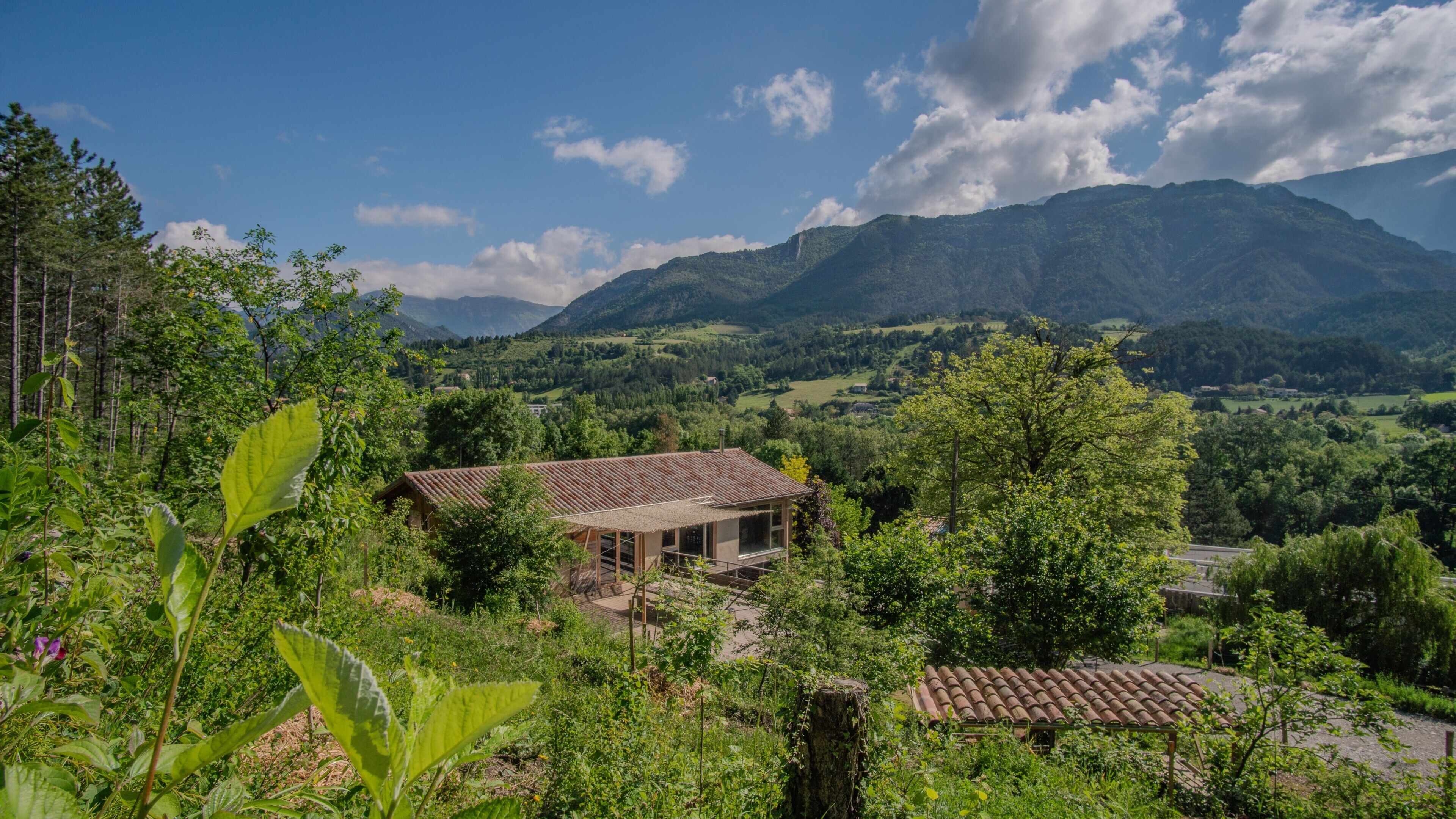 A rustic house with a tiled roof, surrounded by lush greenery and mountains in the background.
