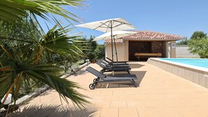 Un espace aménagé au bord de la piscine, avec des chaises longues et des parasols, un bâtiment doté d’un toit en tuiles et une piscine.