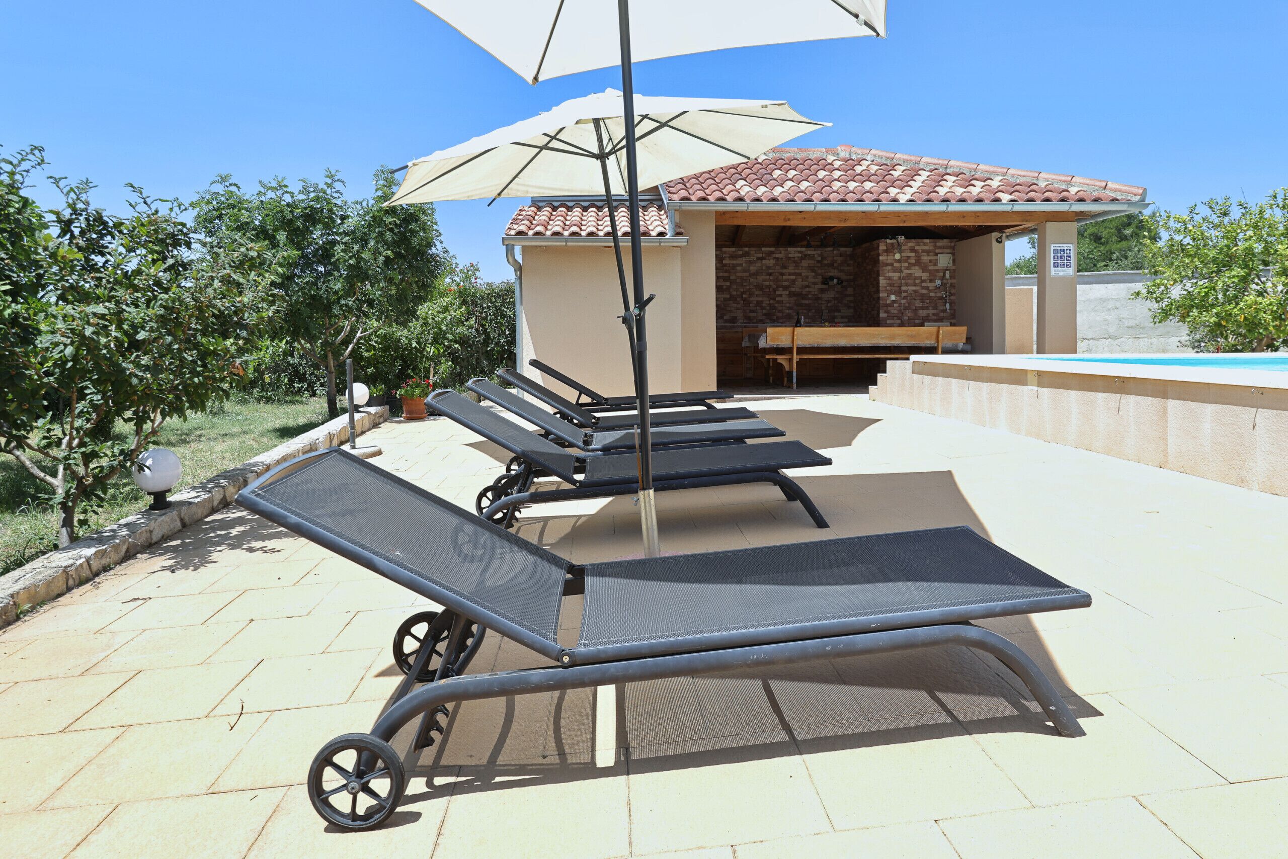 Outdoor lounge chairs, a pergola with an umbrella, and a poolside area with a stone wall and a wooden bench.