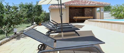 Outdoor lounge chairs, a pergola with an umbrella, and a poolside area with a stone wall and a wooden bench.