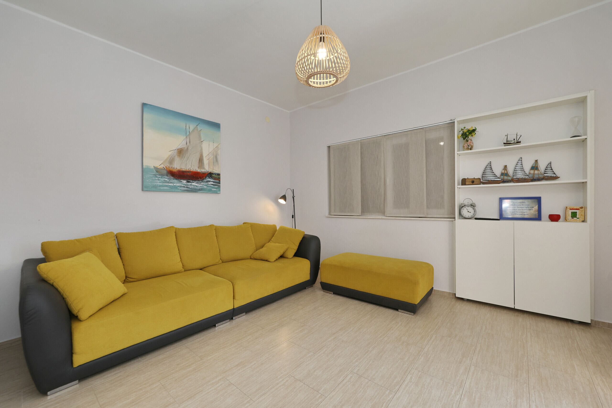 Un salon comprenant un canapé, un pouf, un tableau représentant un voilier, un meuble blanc orné d’objets décoratifs et un luminaire suspendu.